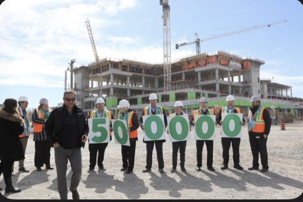 A group in hard hats and safety vests holding signs spelling out 500000 in front of the Niagara Health hospital construction site